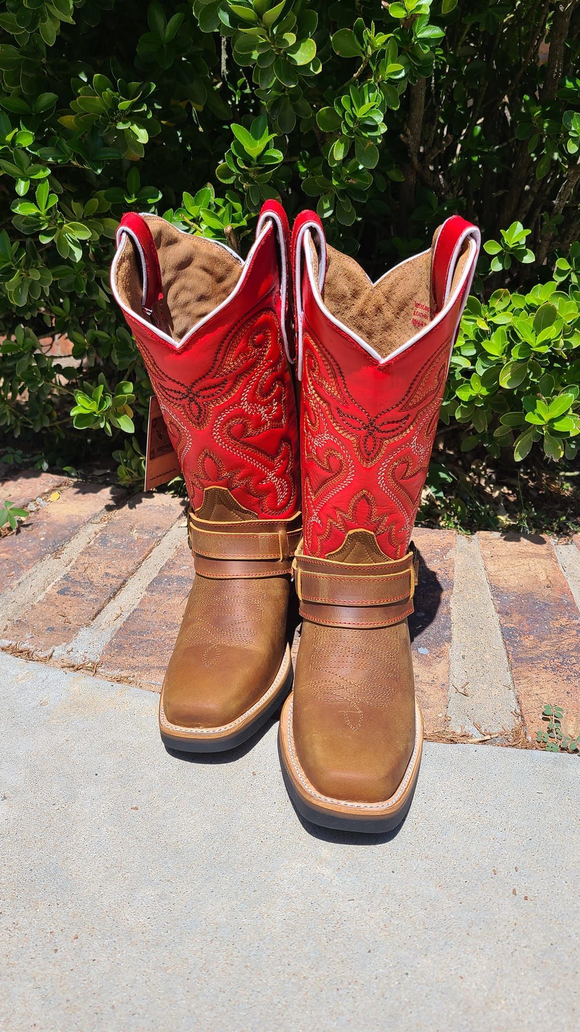 A pair of red and brown cowboy boots are sitting on a sidewalk.