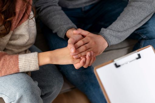 שני אנשים מחזיקים ידיים ברגישות, ברקע לוח כתיבה – סצנה של תמיכה רגשית או שיחת טיפול