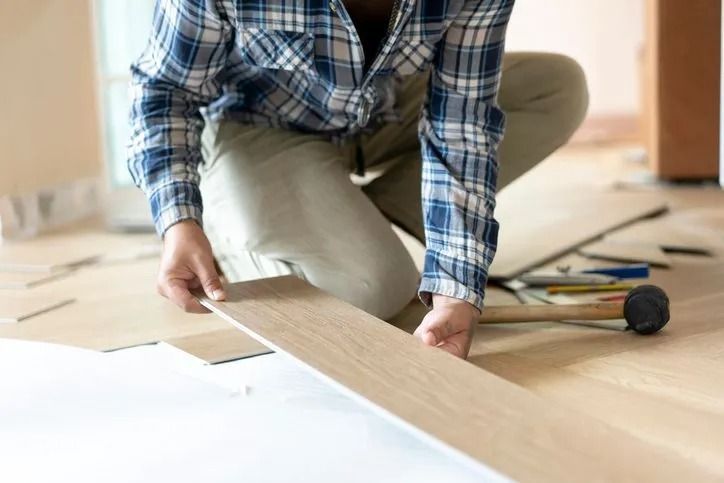 Persona che installa pavimenti in legno, inginocchiata, con camicia a quadri e pantaloni beige, in un ambiente interno.