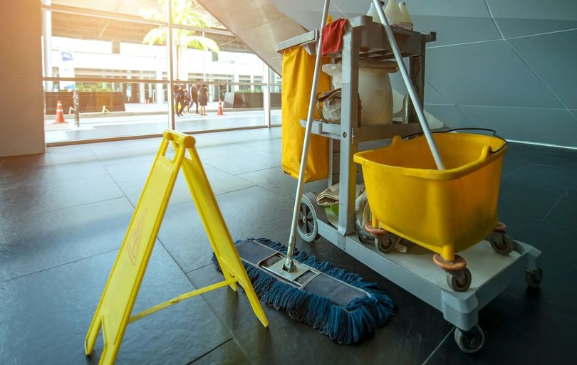 Cartello giallo di pavimento bagnato e carrello per la pulizia con mocio in un edificio con ampie finestre.