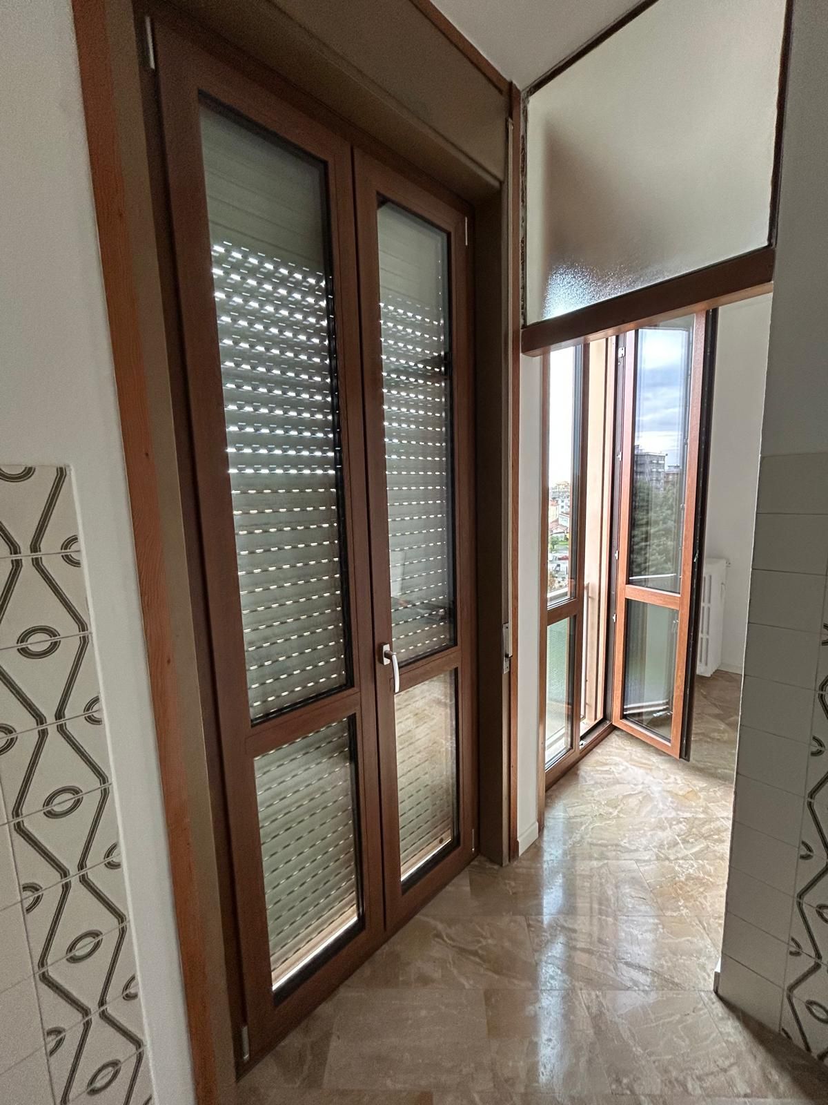 Un piccolo balcone con finestra con cornice in legno, tenda avvolgibile e vista sugli alberi.