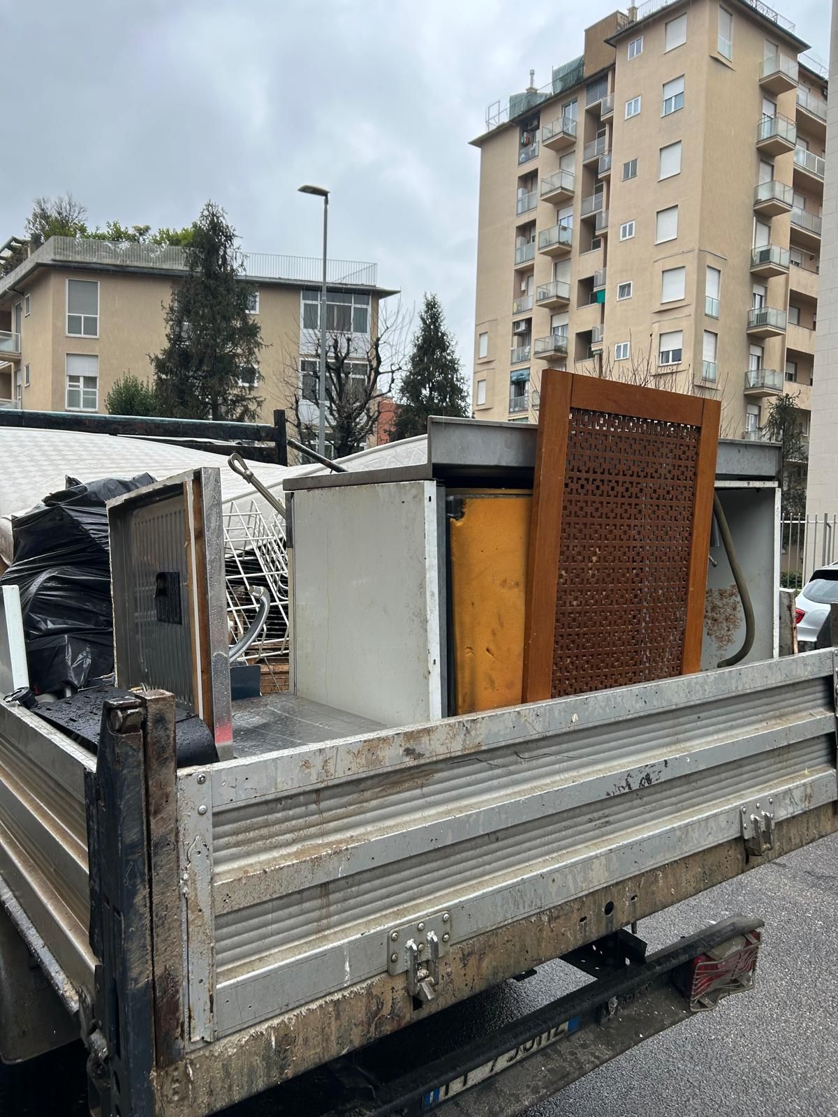 Il cassone di un camion pieno di metallo, legno e detriti di fronte agli edifici sotto un cielo coperto.