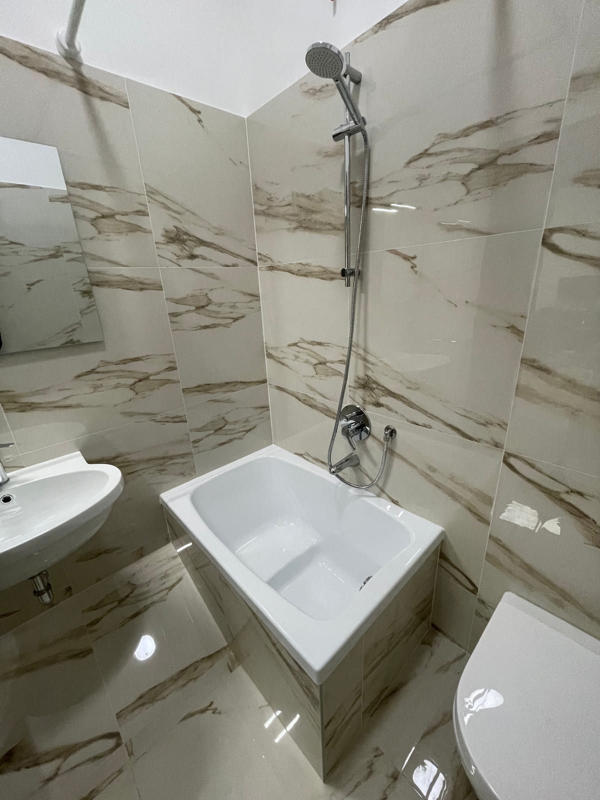 Bagno con sanitari bianchi, tra cui vasca, lavandino e WC, in contrasto con pareti e pavimento piastrellati beige e marroni effetto marmo.