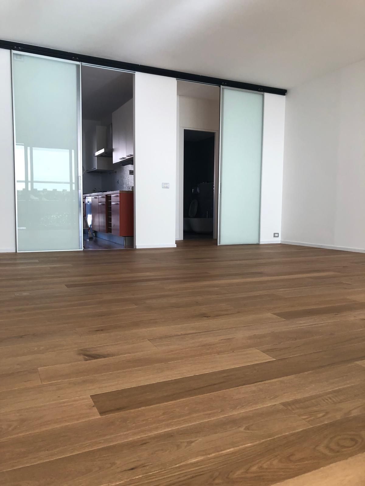 Pavimento in legno in una stanza con porte scorrevoli in vetro smerigliato, che conducono alla cucina e al corridoio buio.