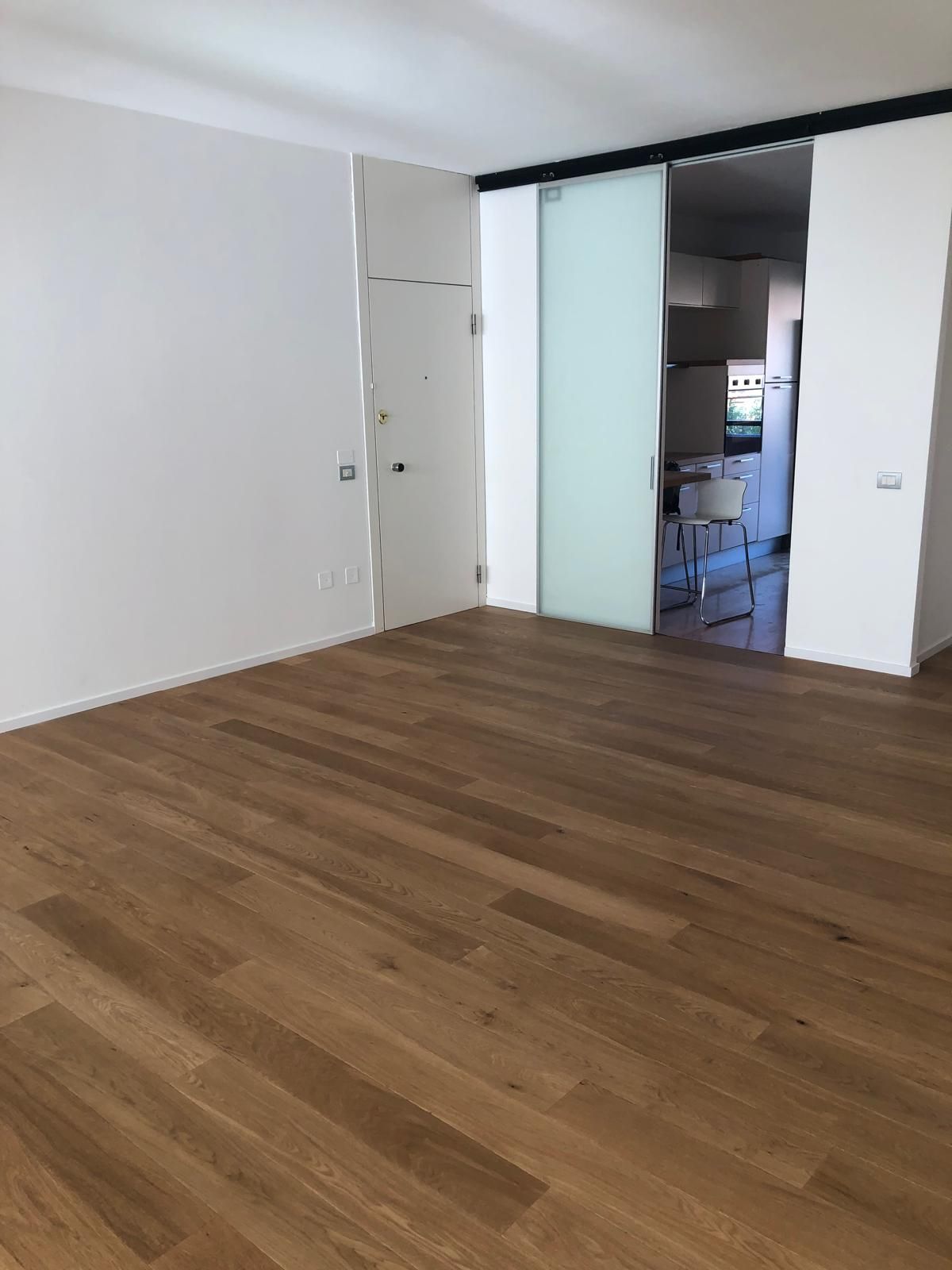 Stanza vuota con pavimento in legno, pareti bianche e una porta scorrevole in vetro smerigliato.
