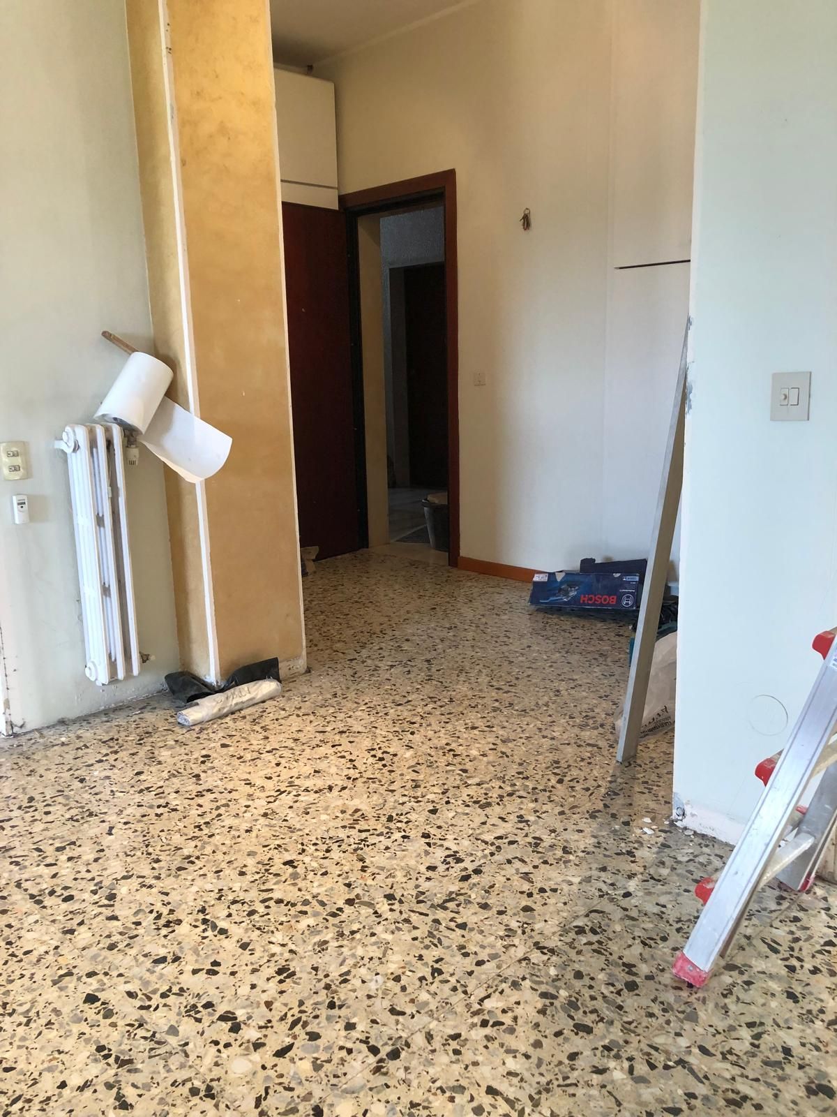 Stanza con pavimento in terrazzo, pareti bianche, porta che conduce a un'altra stanza, un termosifone e una scala.