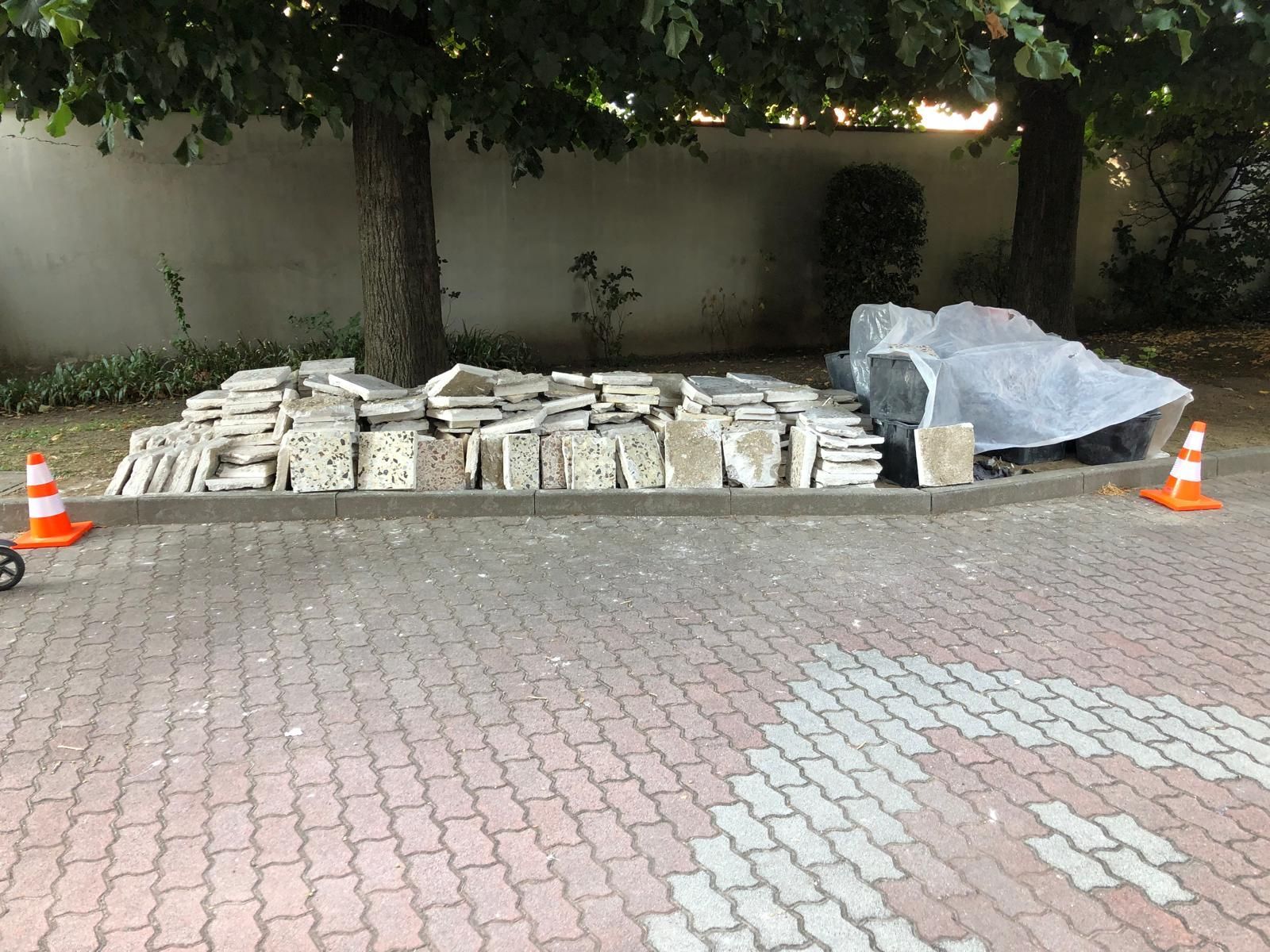 Mucchi di pietre grigie e lastre di pietra coperte su un vialetto di mattoni rossi, vicino ad alberi e coni stradali.