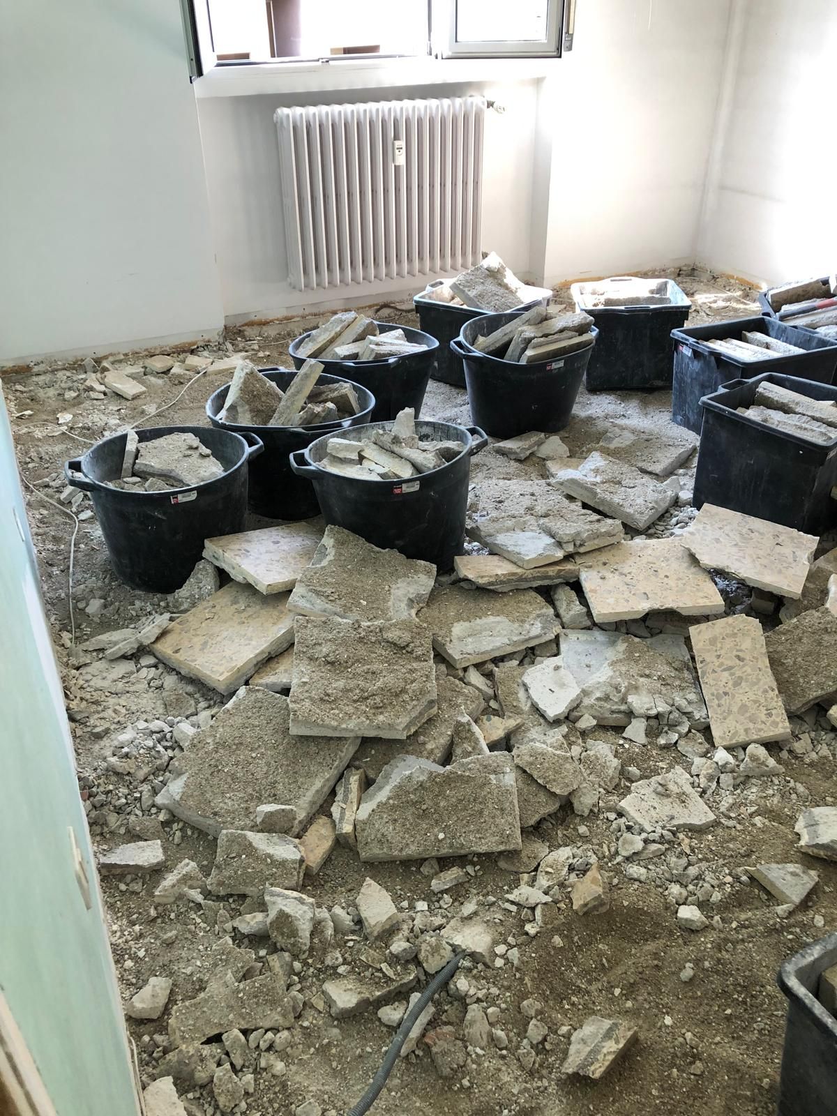 Stanza in fase di ristrutturazione. Detriti di cemento e secchi pieni di piastrelle rotte su un pavimento polveroso.