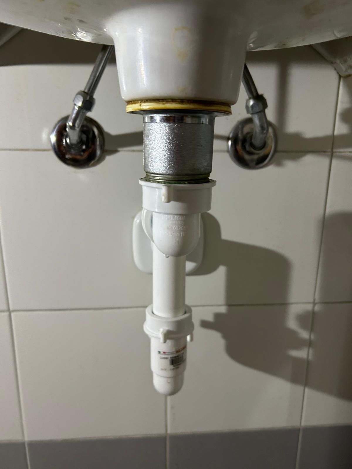 Parte inferiore di un lavandino bianco con tubi di scarico collegati, appoggiato a una parete piastrellata bianca.