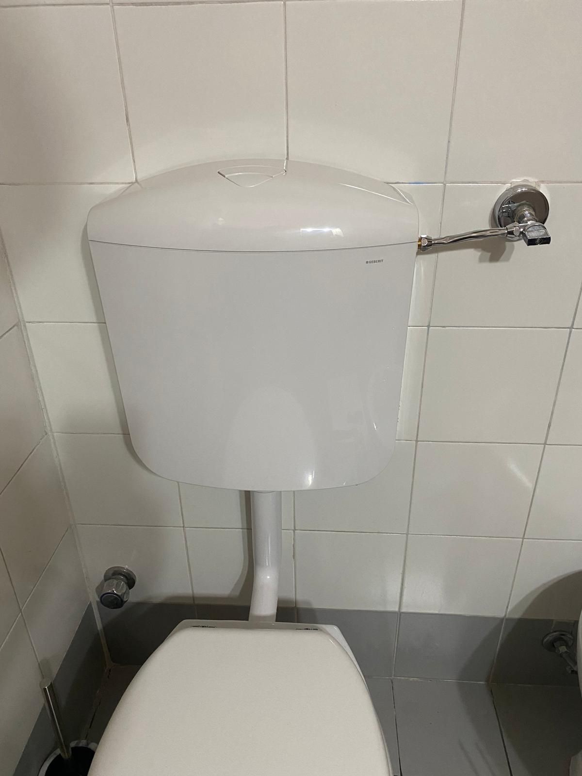 Cassetta del water bianca su parete piastrellata bianca. Leva di scarico cromata, sedile del water visibile.