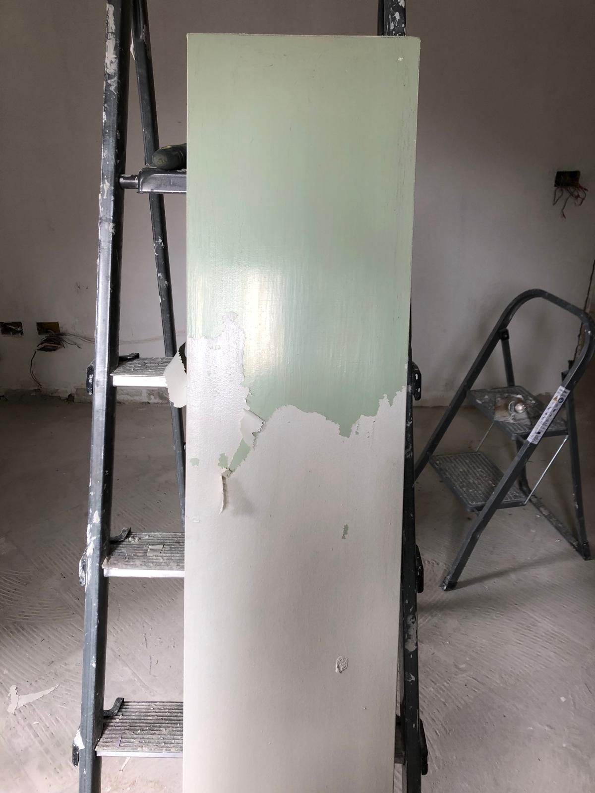 Una porta dipinta a metà di verde chiaro, con la vernice che si stacca dalla parte non verniciata. Sullo sfondo, una scala e uno sgabello.