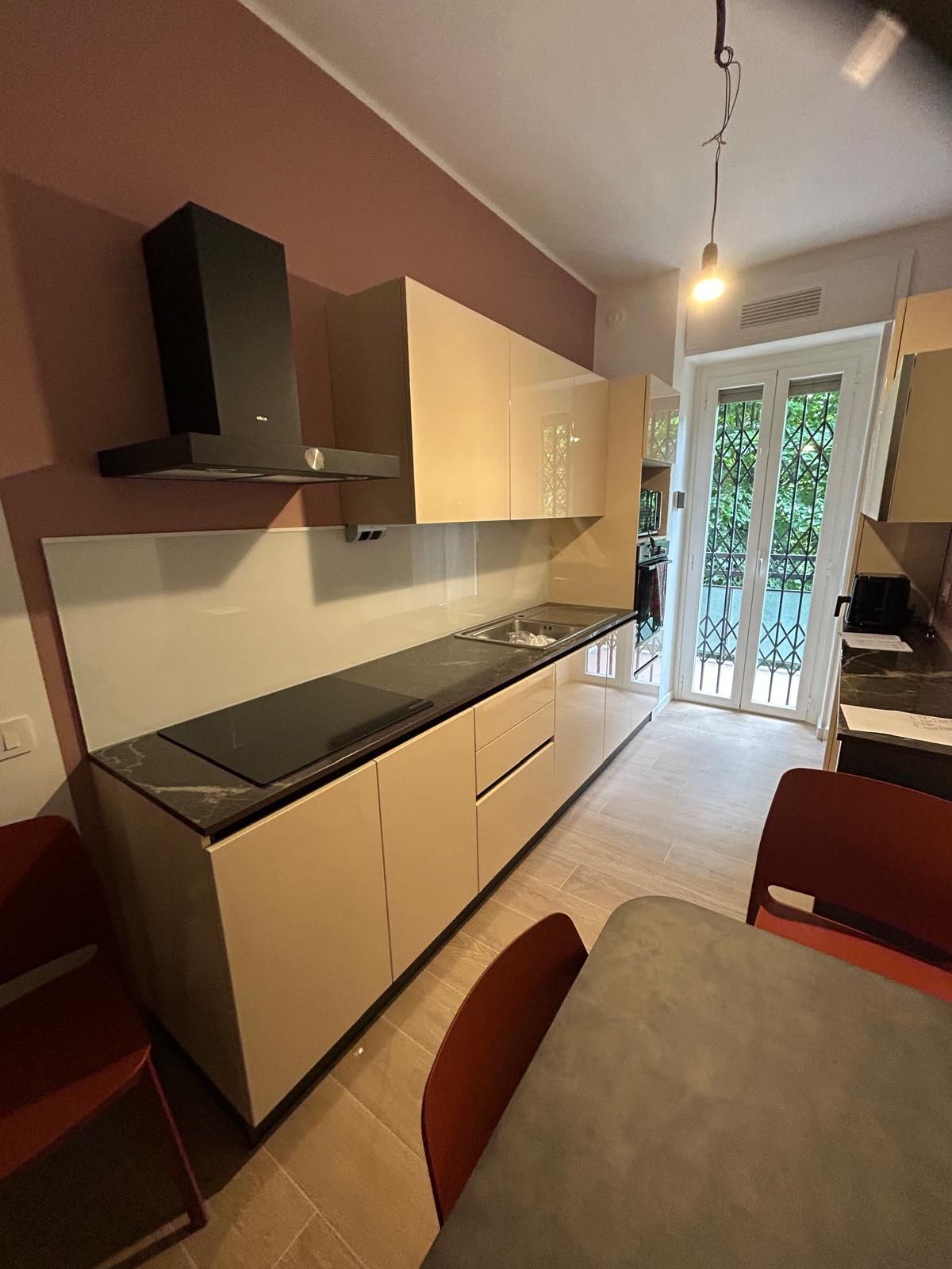 Cucina moderna con mobili beige, elettrodomestici neri, sedie rosse e balcone con vista.