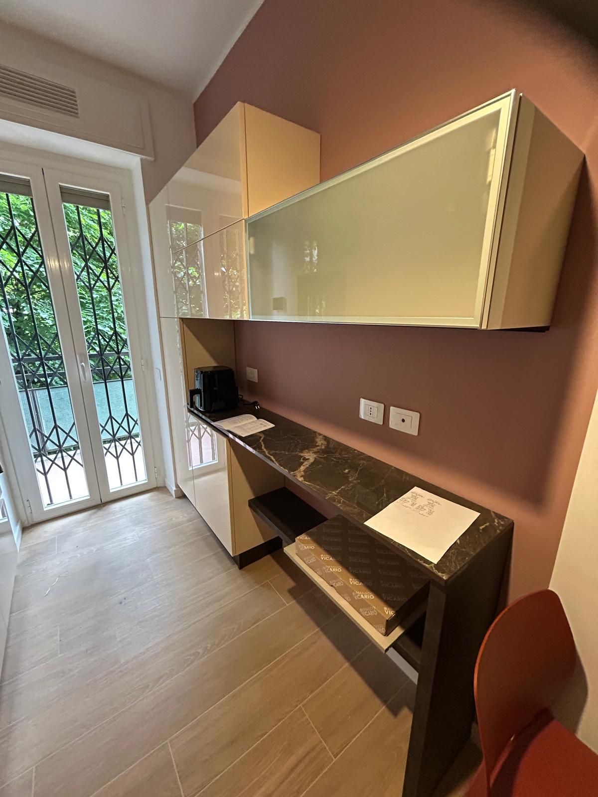 Scrivania con piano in marmo scuro, mobili color crema e parete color cipria. Sulla sinistra si trova una porta che dà su un balcone.