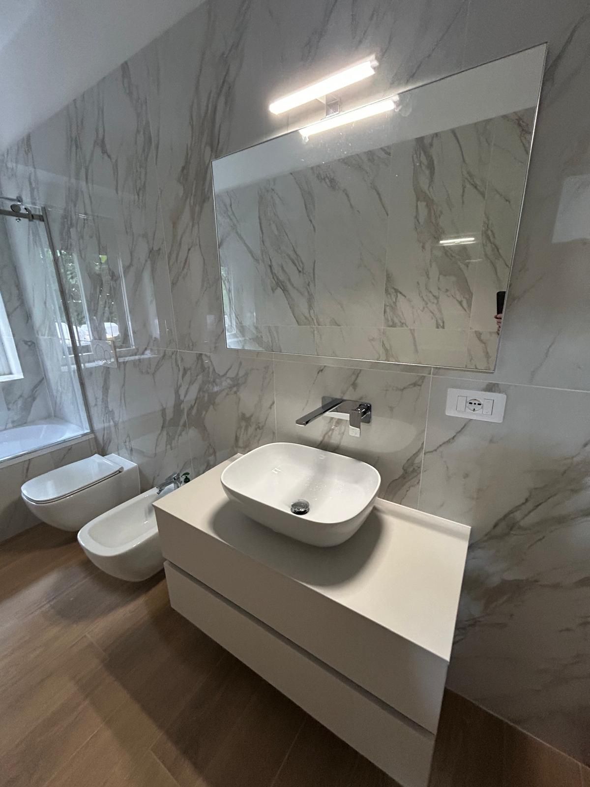 Bagno moderno con sanitari bianchi, pareti con motivi in ​​marmo e un grande specchio.