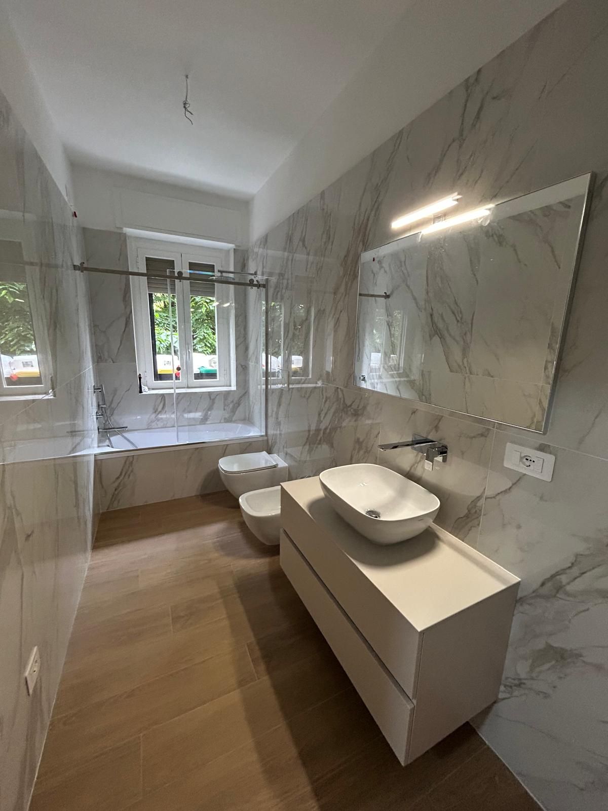 Bagno con pareti effetto marmo, sanitari bianchi e pavimento in legno. Dotato di lavabo, WC, bidet e vasca.