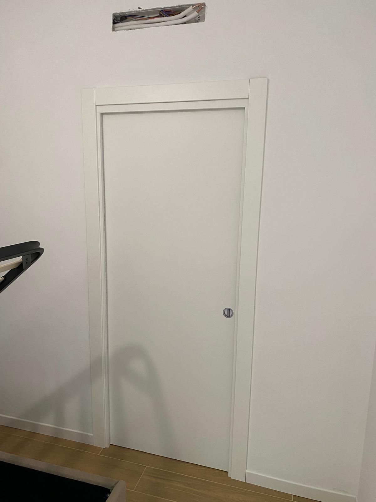 Porta bianca con telaio in una stanza dai colori chiari, accanto a una parete e pavimento in legno.