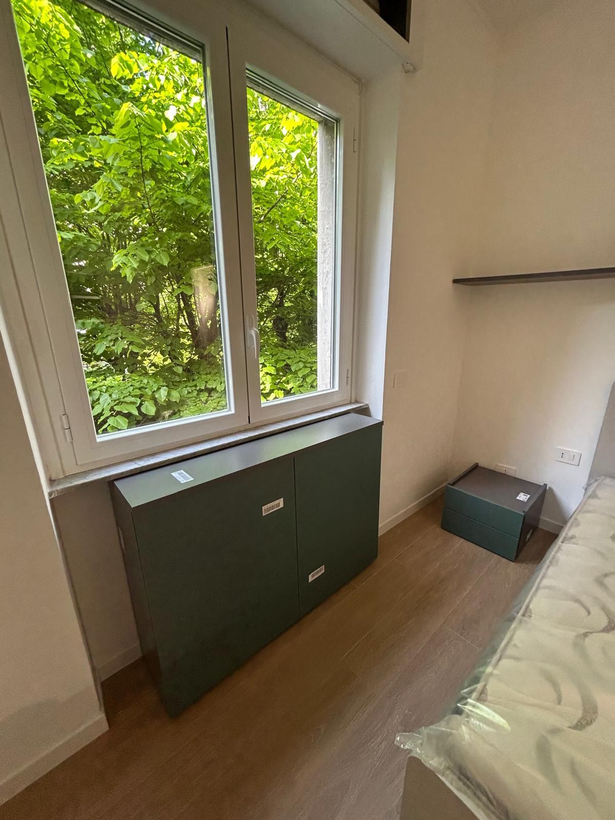 Una stanza con una finestra che si affaccia sul verde. Sotto la finestra c'è un mobiletto verde e sul pavimento c'è una piccola scatola.
