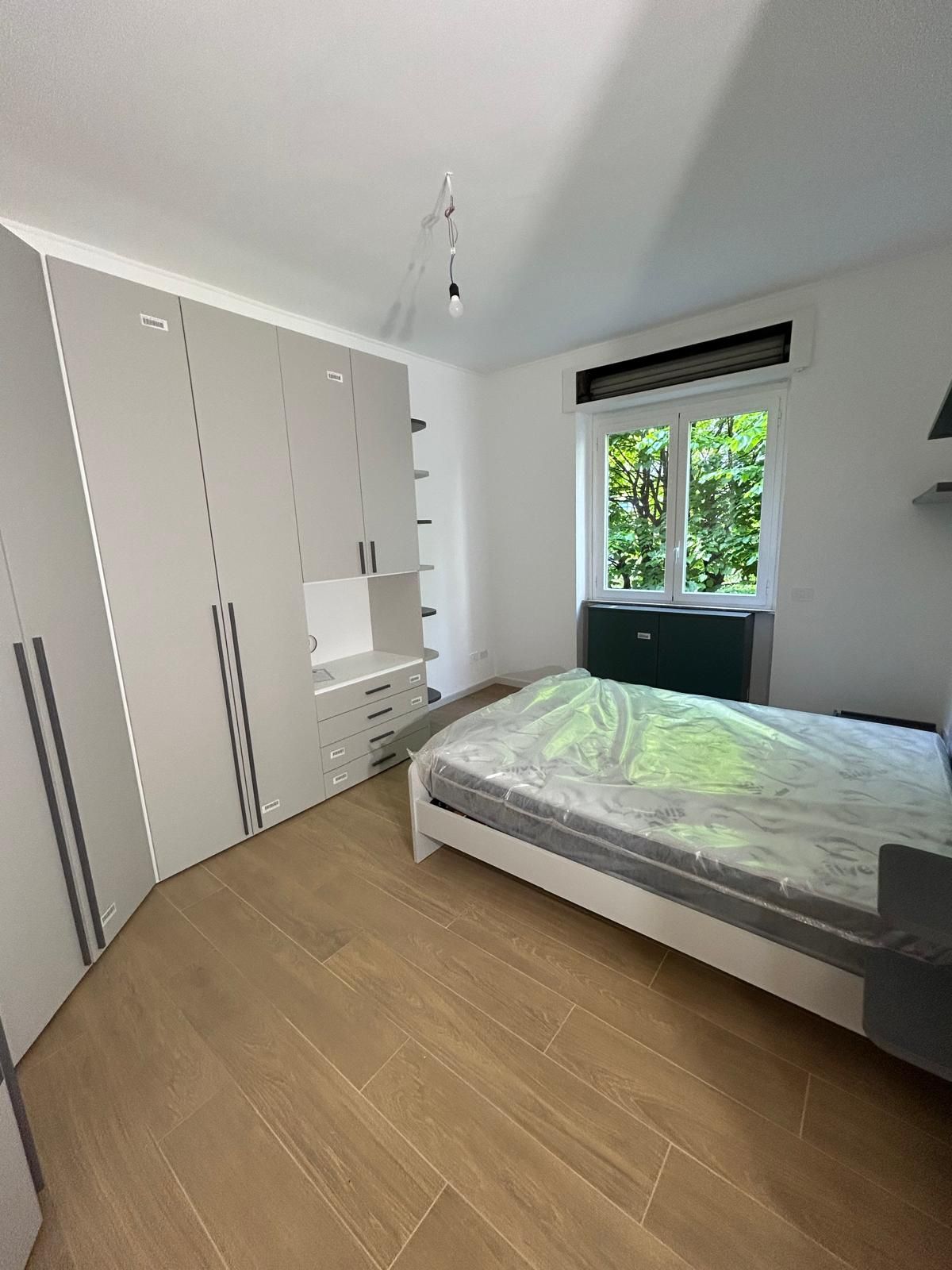 Camera da letto con pavimenti in legno chiaro, grande armadio bianco, letto e finestra con vista sull'esterno.