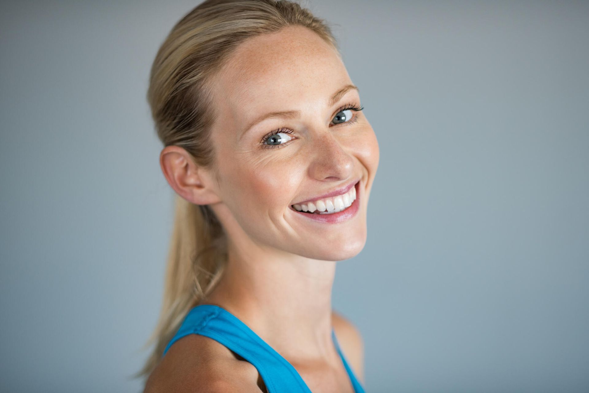 Woman with blonde ponytail, smiling, wearing a blue top, against a gray background.