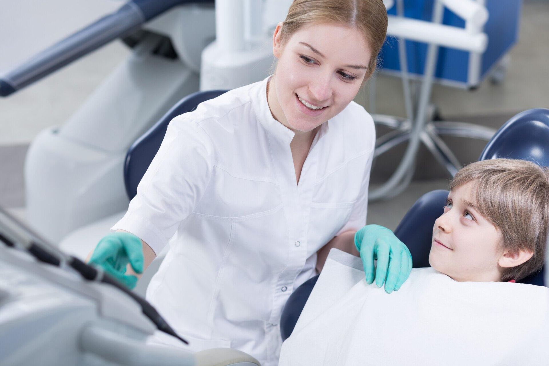 A female dentist is talking to a young boy in a dental chair.