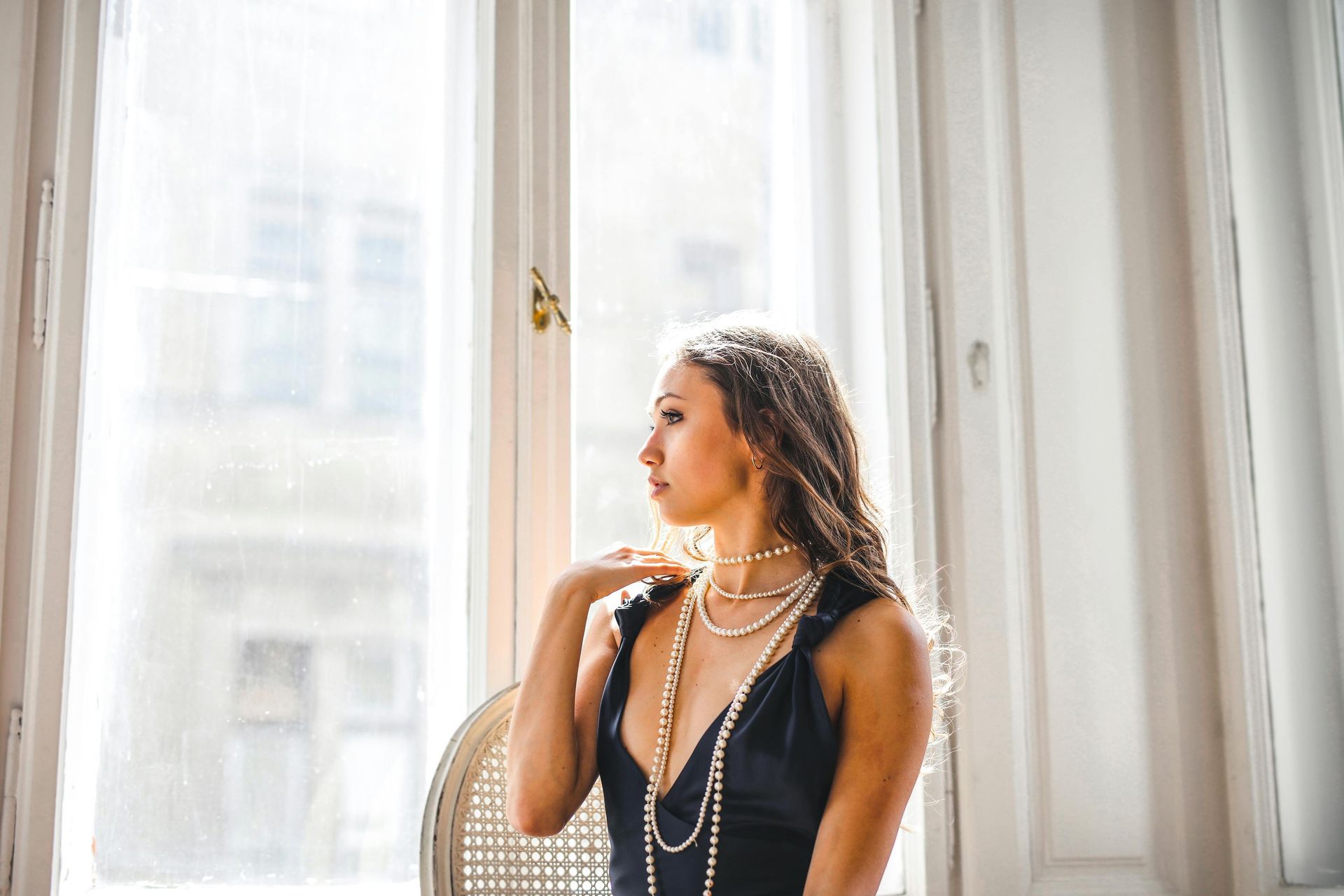 Woman looking out of a window dressed up with a black dress and layered necklaces