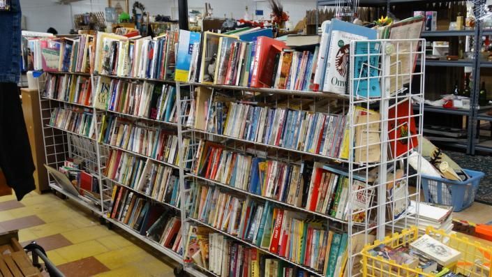 A crowded used bookstore aisle with leaning shelves packed with colorful books and DVDs