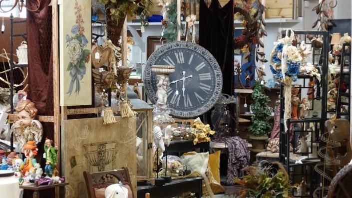 Cluttered antique shop with a large gray clock, hanging decor, and shelves of vintage items