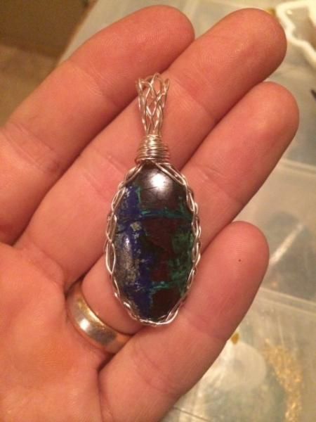 Hand holding an oval blue-green iridescent pendant with a silver wire wrap and chain bail