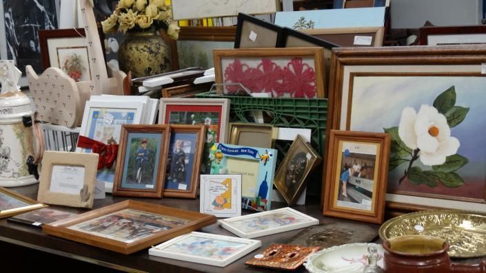 Assorted framed artwork and decorative dishes displayed on a crowded table in a shop or market stall