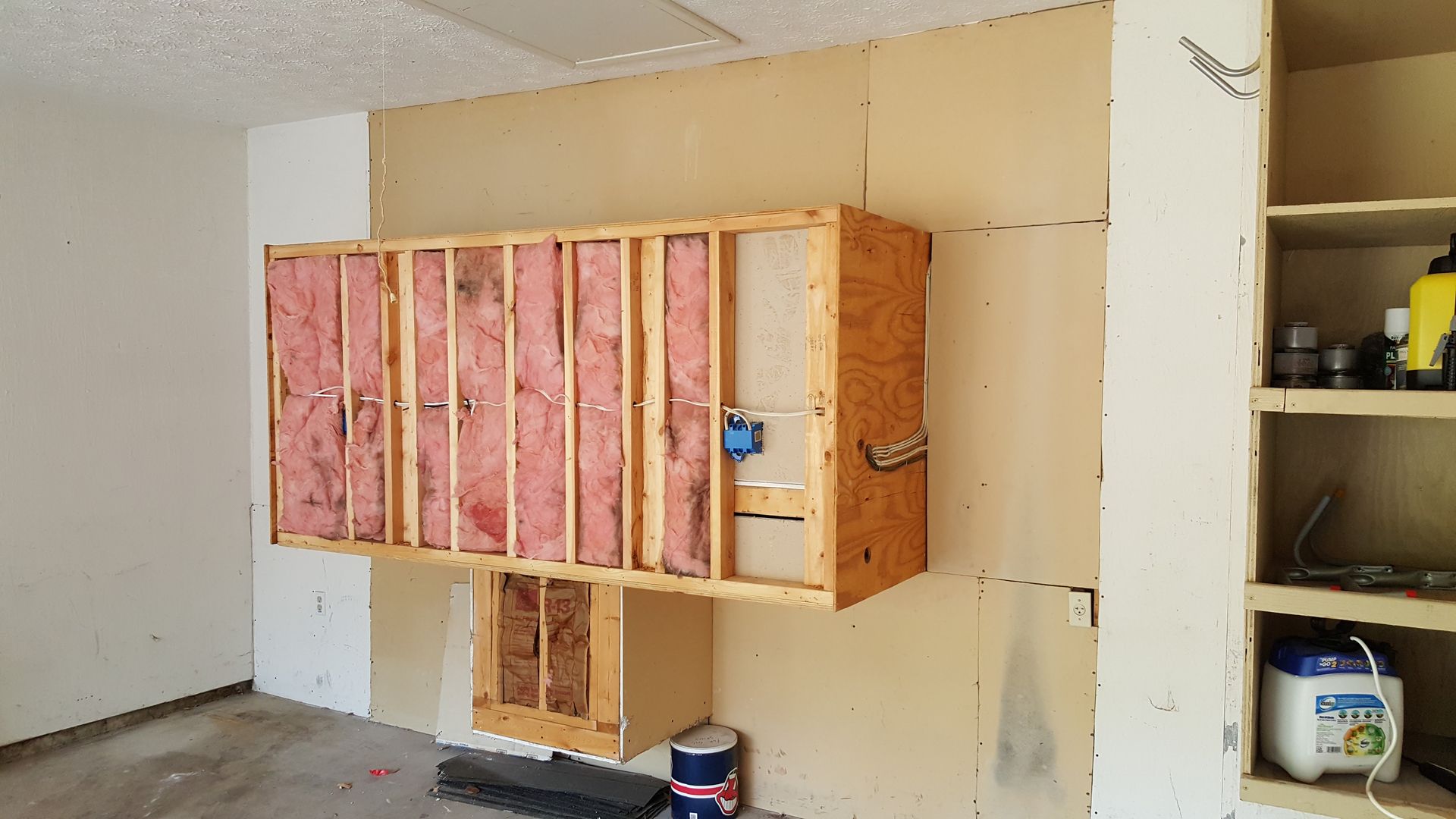 A room with a wooden frame on the wall and a bucket of laundry detergent.