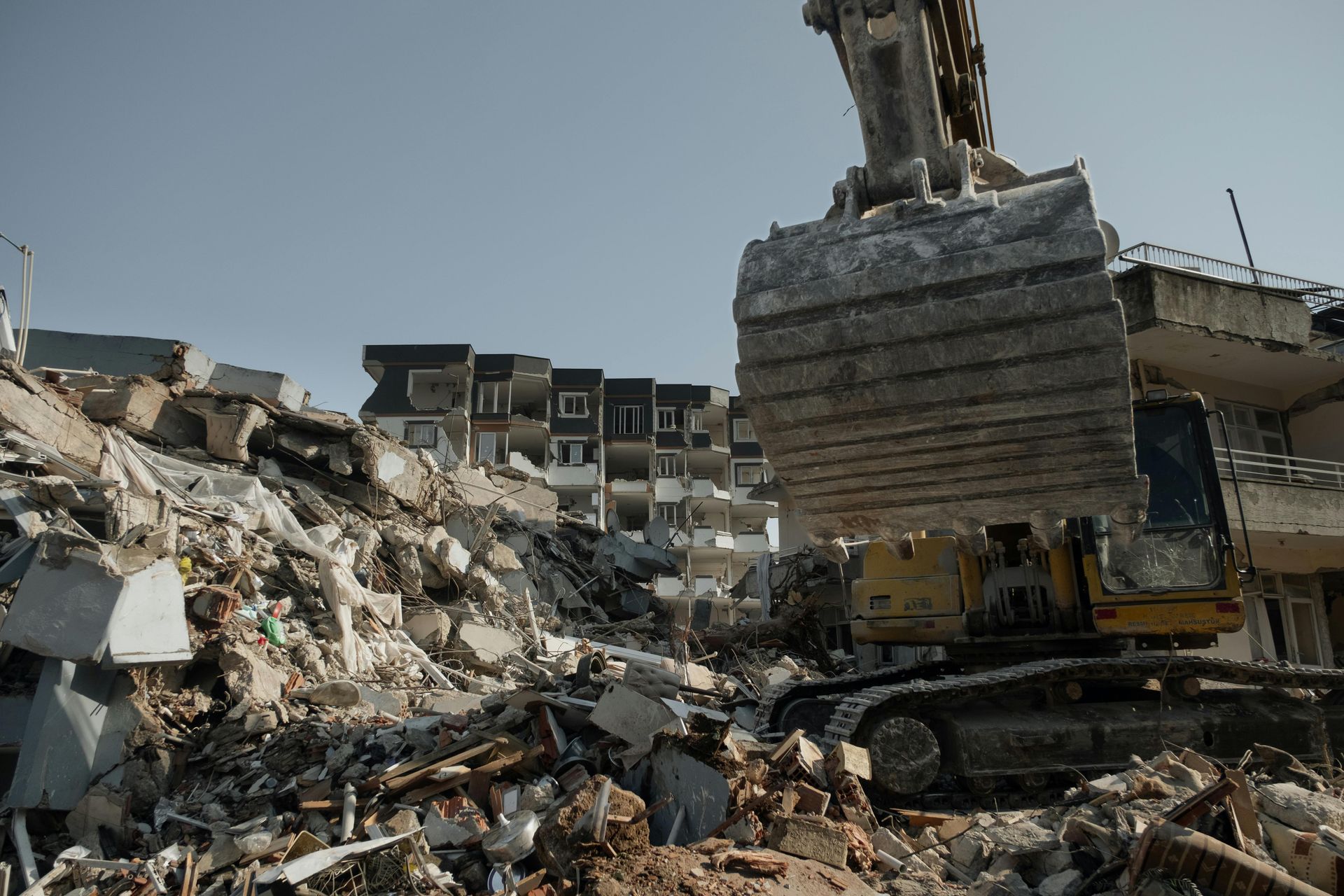 Excavator demolishing debris of a collapsed building with a partially damaged structure in the background.