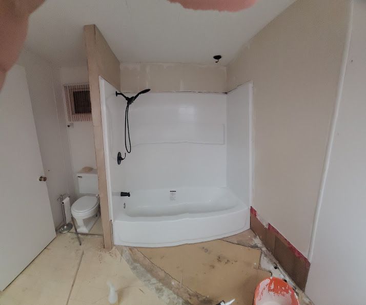 Bathroom remodel in progress: white tub/shower, black showerhead, toilet, partially finished walls, debris on the floor.