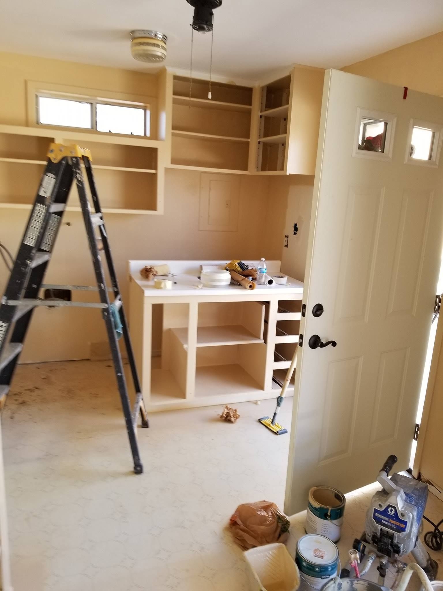 Interior of a room under construction. Cabinets, a door, a ladder, and supplies are visible.