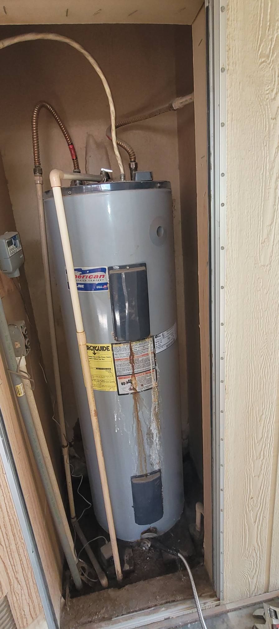 A water heater is housed in a narrow, wooden compartment. It is grey, with pipes and electrical cords visible.