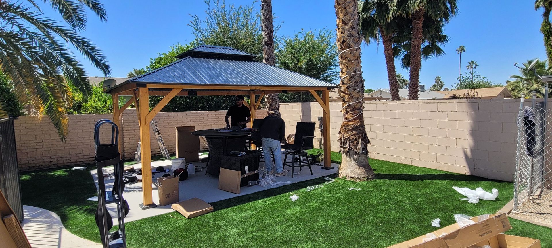 Two people assembling a gazebo in a backyard with green grass and palm trees.