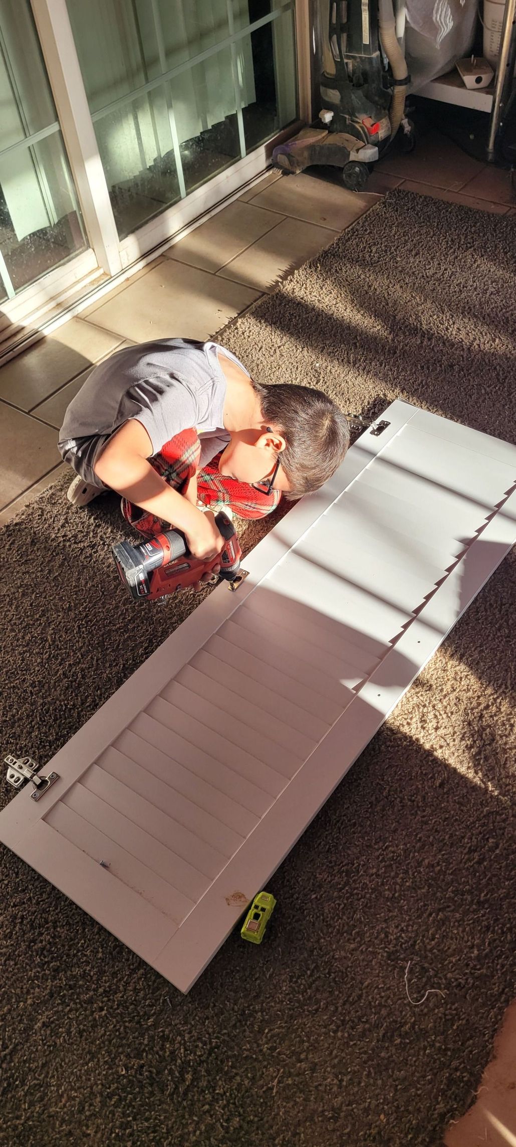 A person leans over a doorway, with a small toy car next to it on a rug in the sunlight.