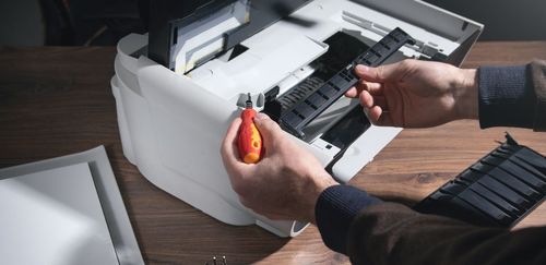 Person repairing a white printer with a screwdriver, on a wooden desk.