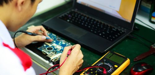 Person repairing a circuit board with a laptop and multimeter on a desk.