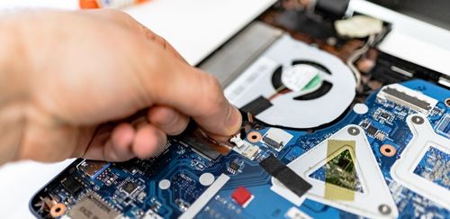 Hand repairing a laptop's circuit board, connecting a cable near a cooling fan.