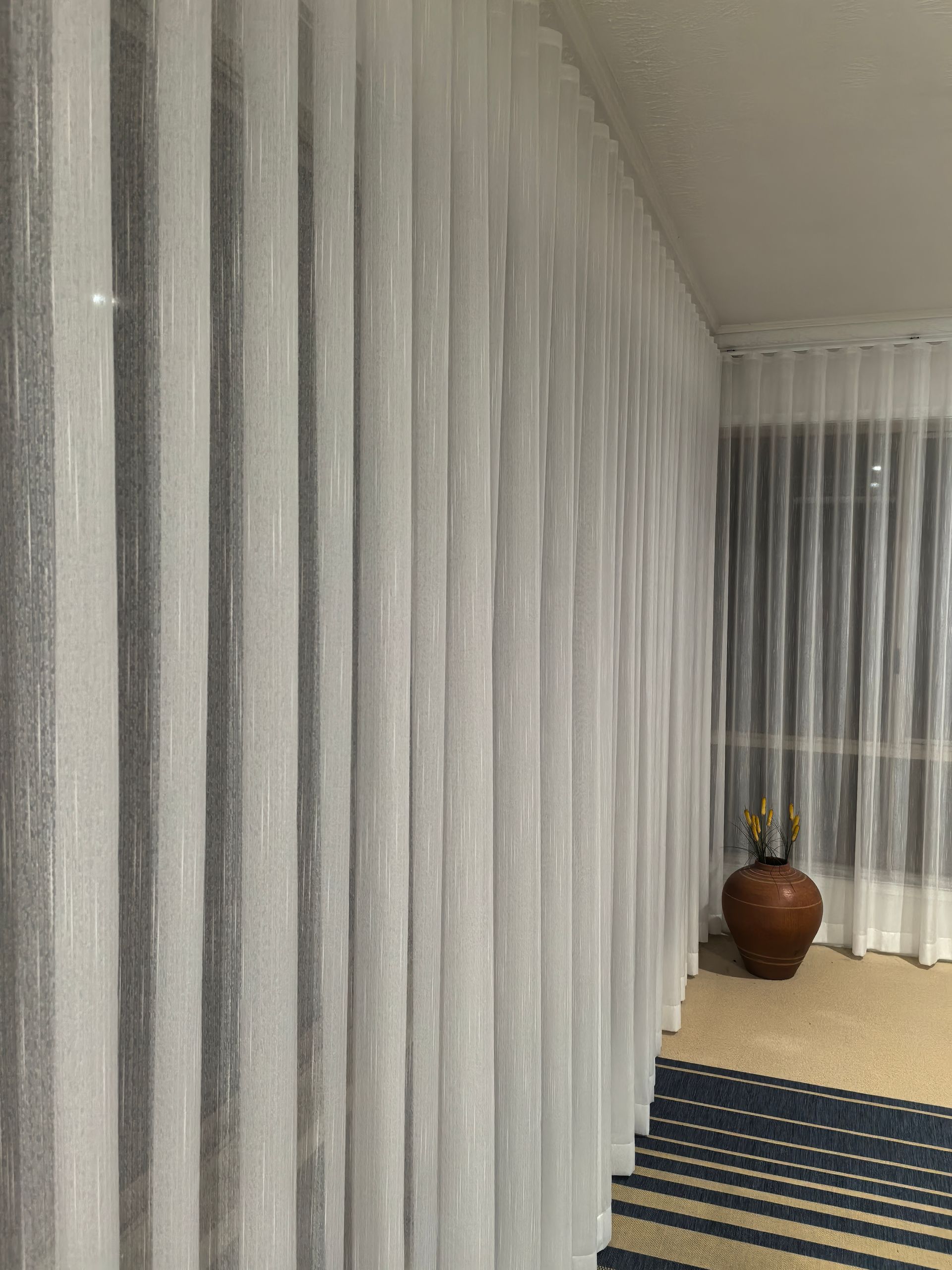 A room with white curtains and a vase on the floor.