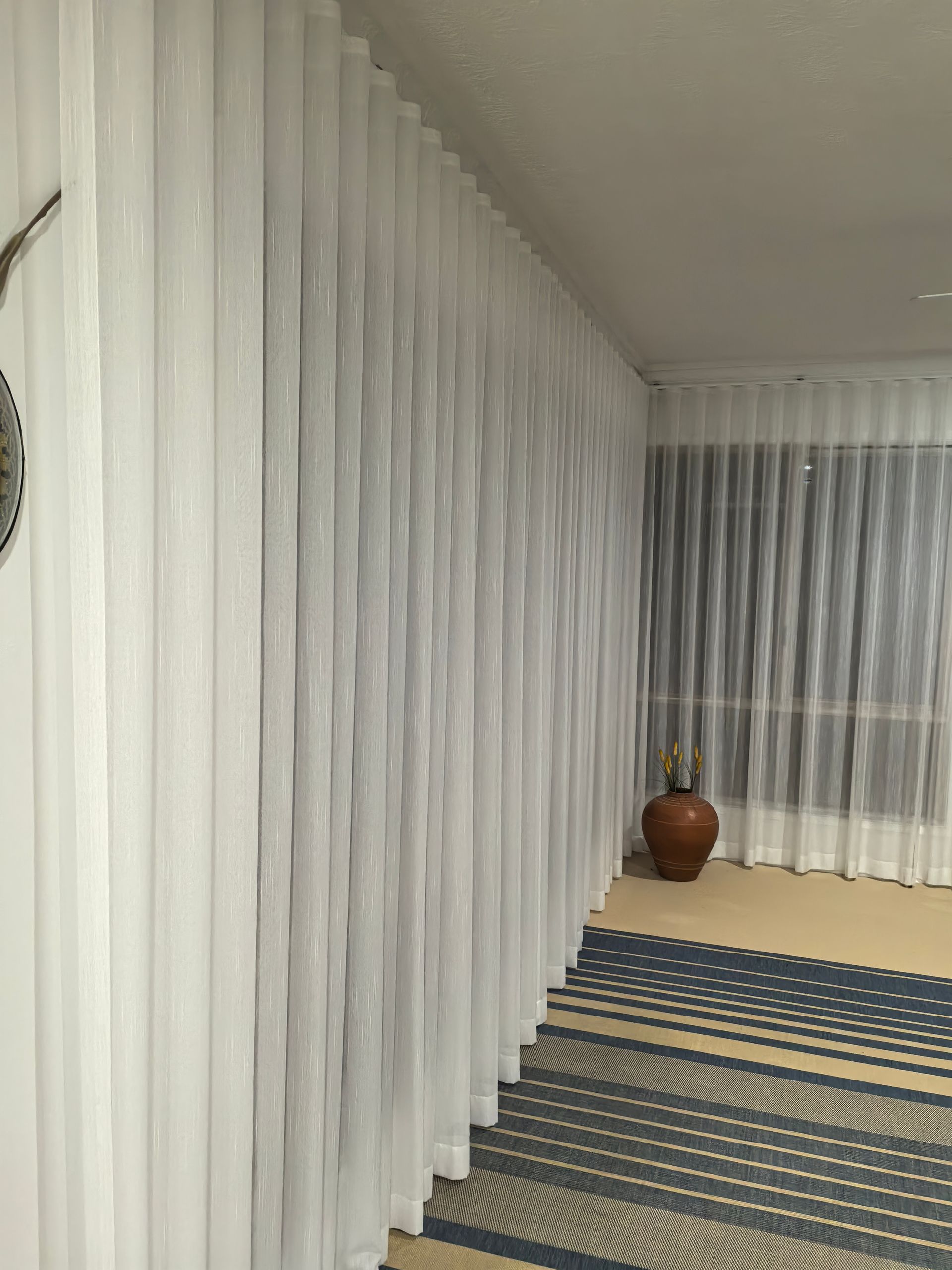 A room with white curtains and a vase on the floor.