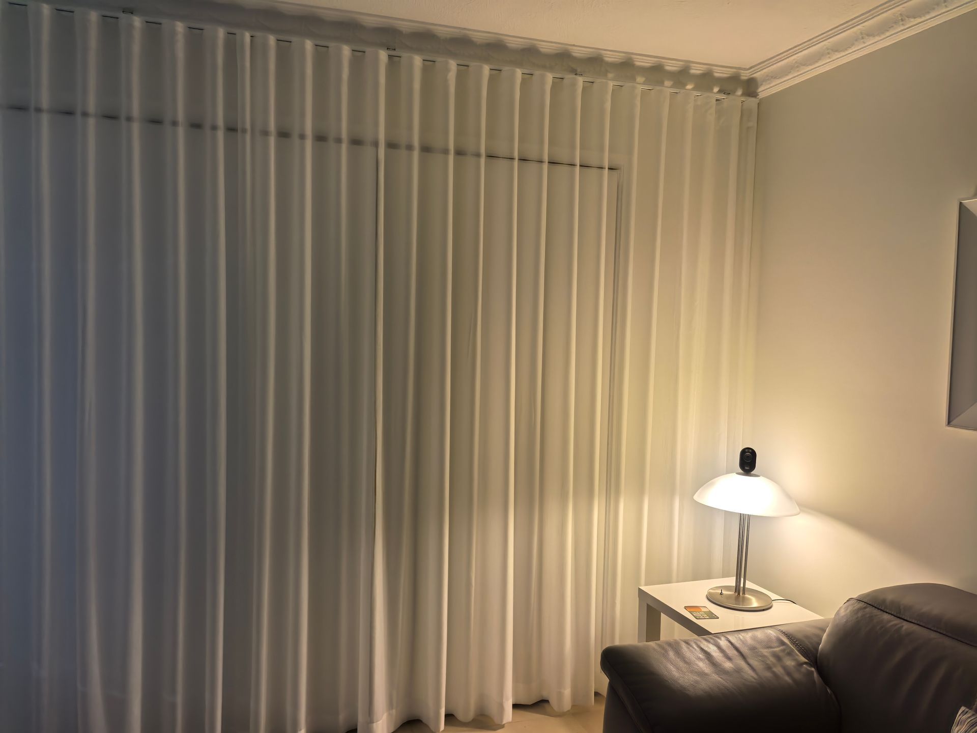 A living room with a couch , table , lamp and white curtains.