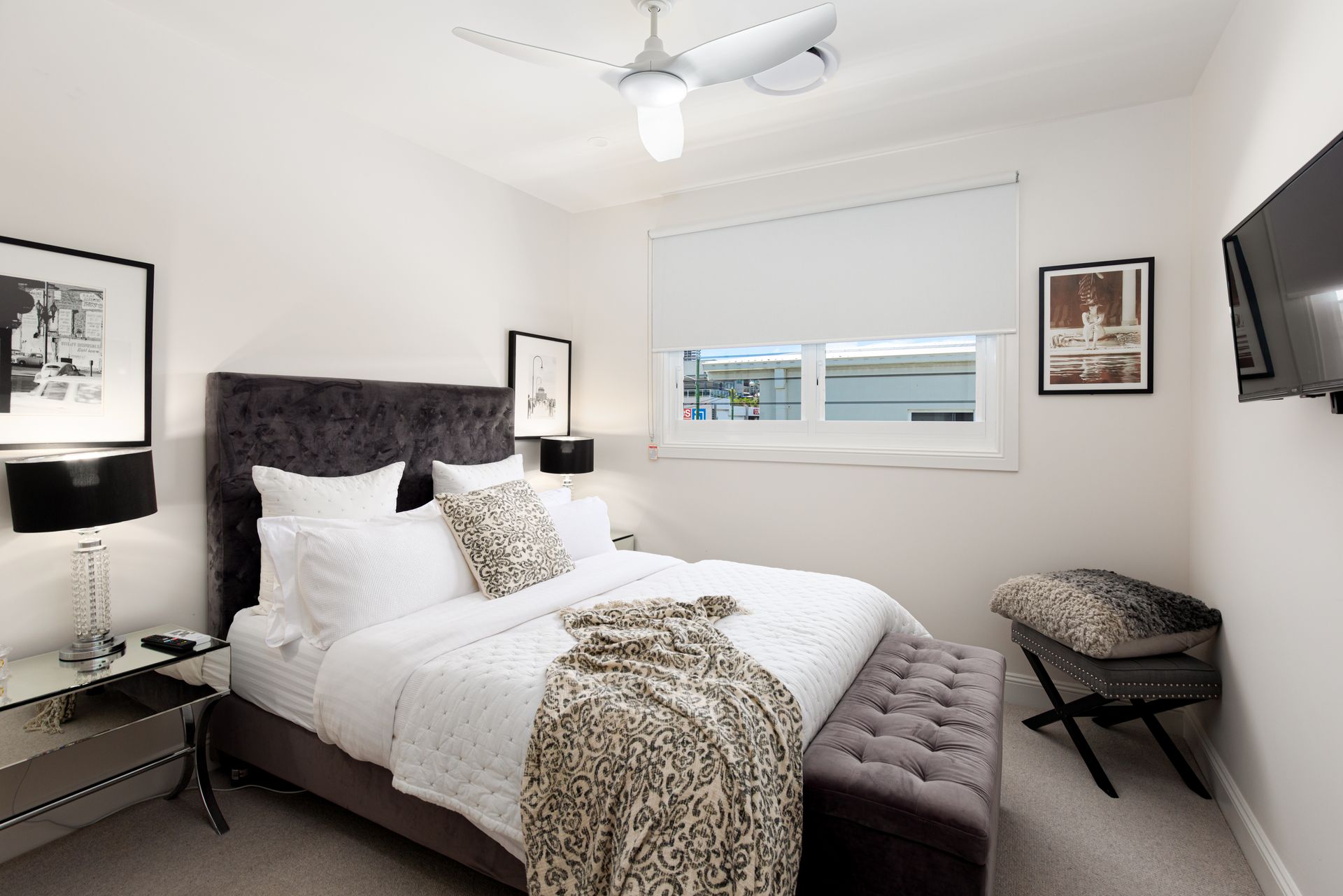 A bedroom with a large bed , a bench , a television and a ceiling fan.