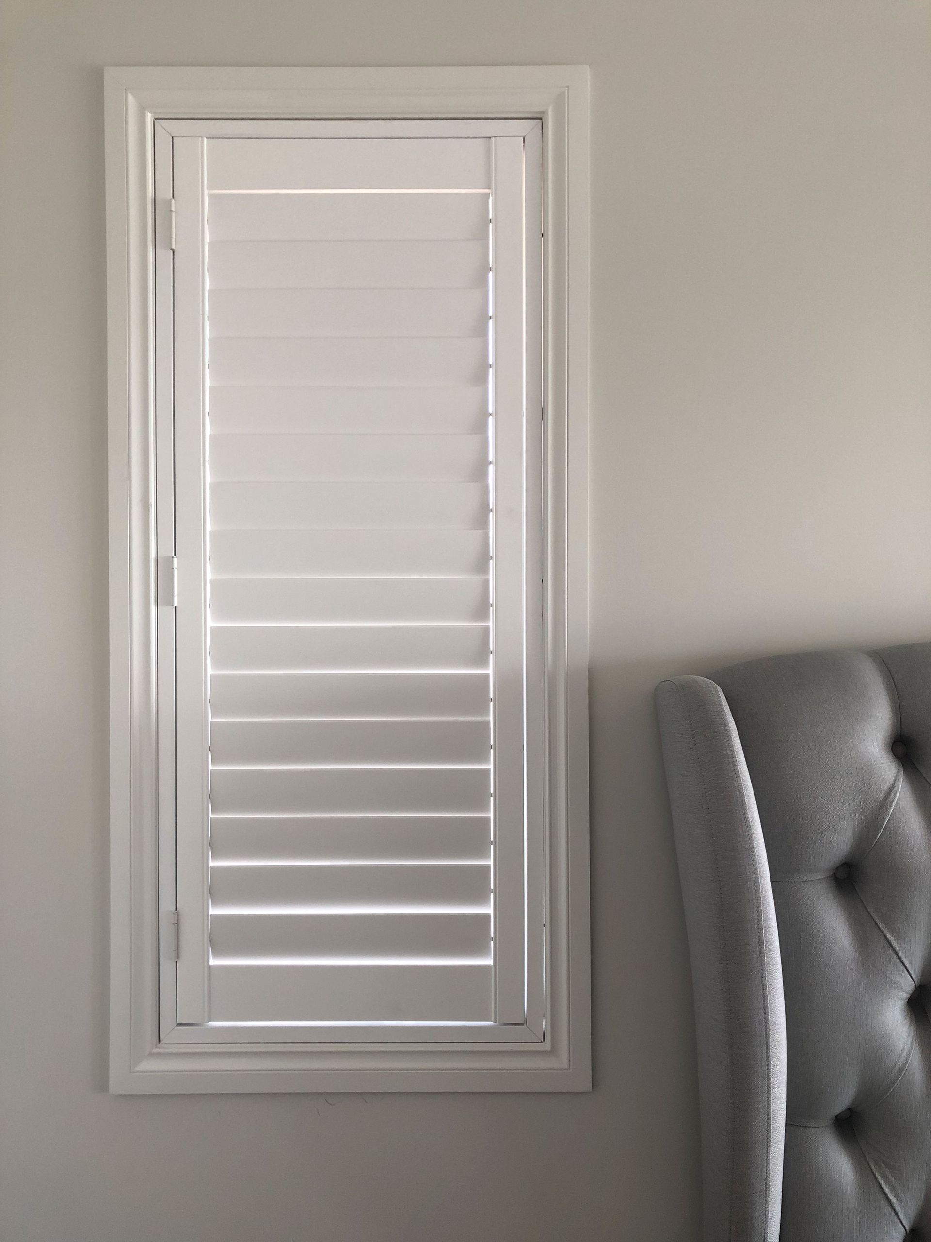 A white shutter is hanging on a wall next to a bed.