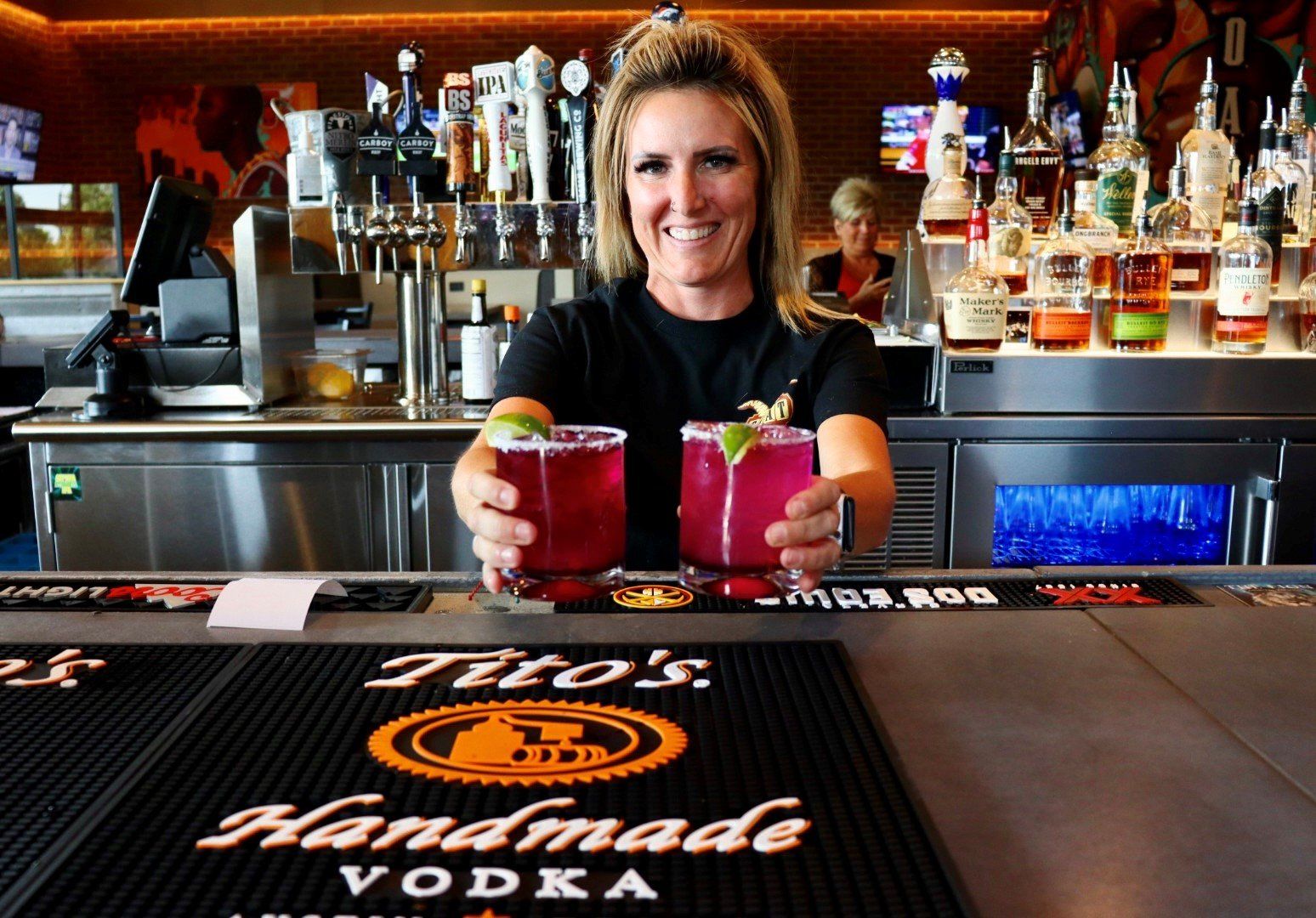 A woman holding two drinks in front of a bar that says tito 's handmade vodka