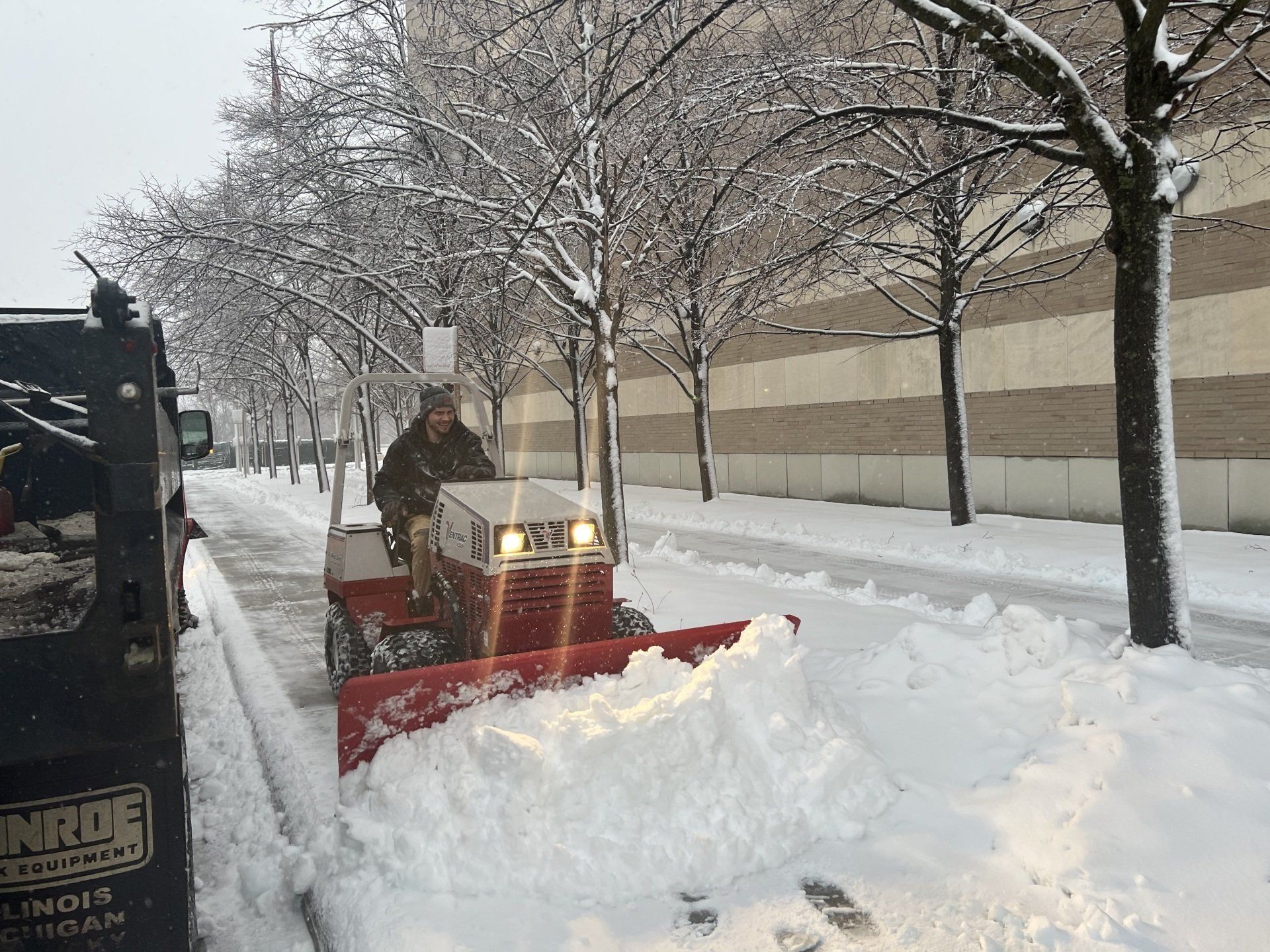 A man is driving a snow plow down a snow covered sidewalk.