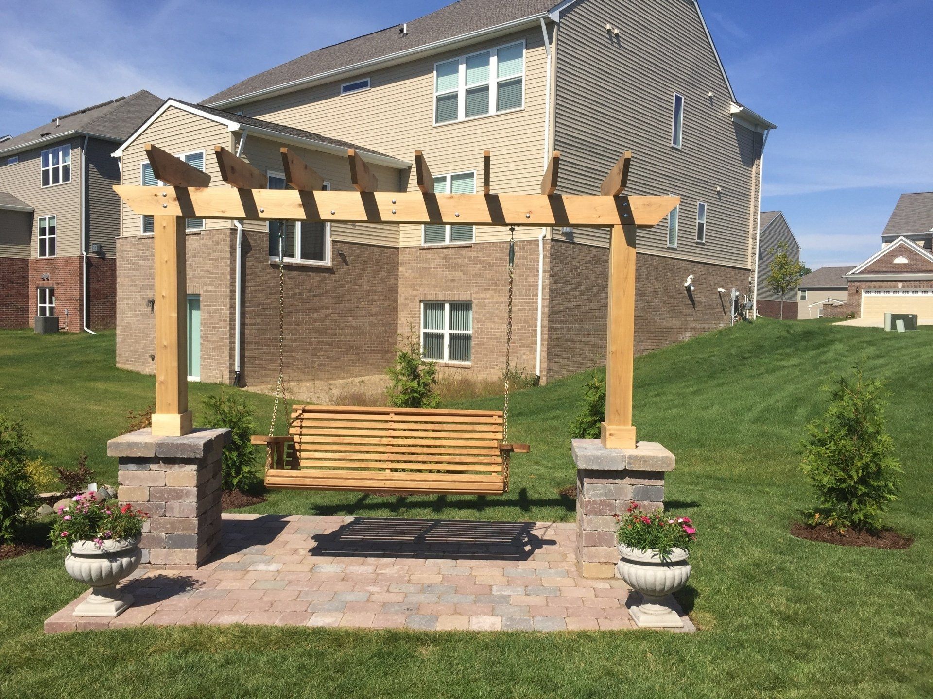 A wooden pergola with a swing underneath it in front of a house.