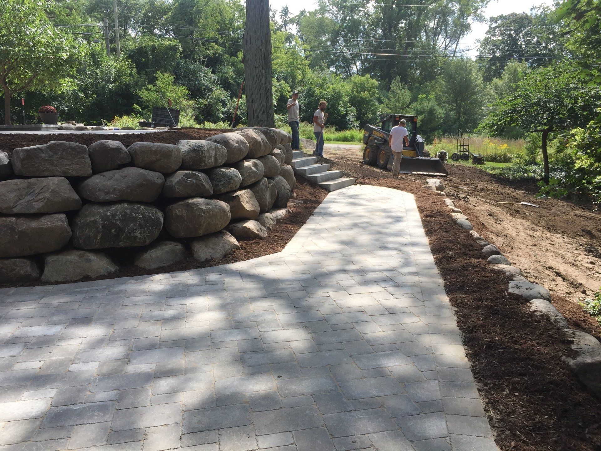 A group of people are standing on a sidewalk next to a stone wall.