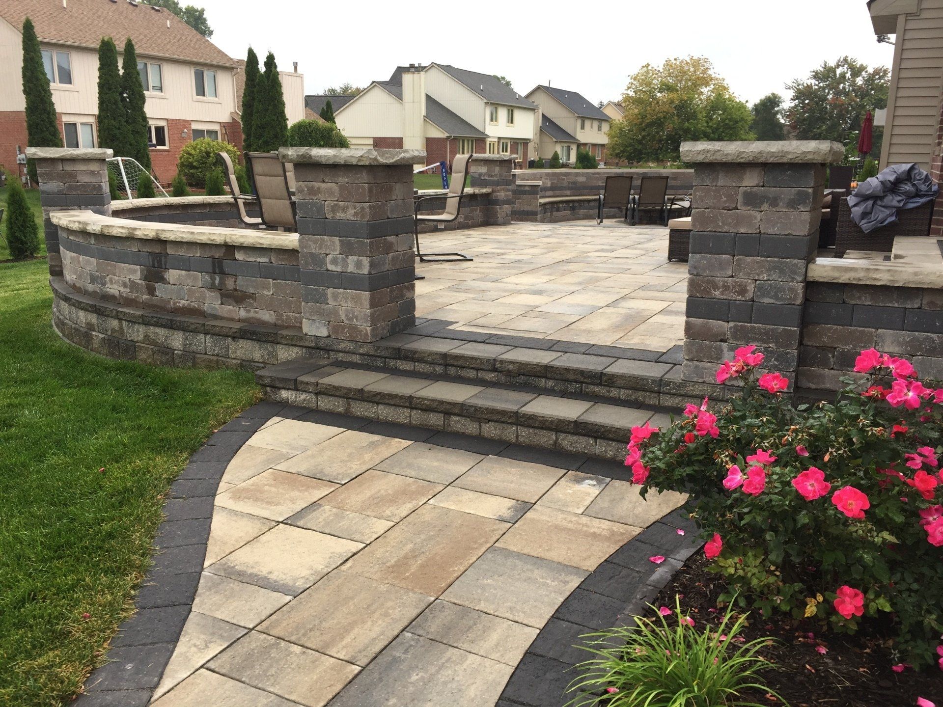 A brick walkway leading to a patio with chairs and flowers.