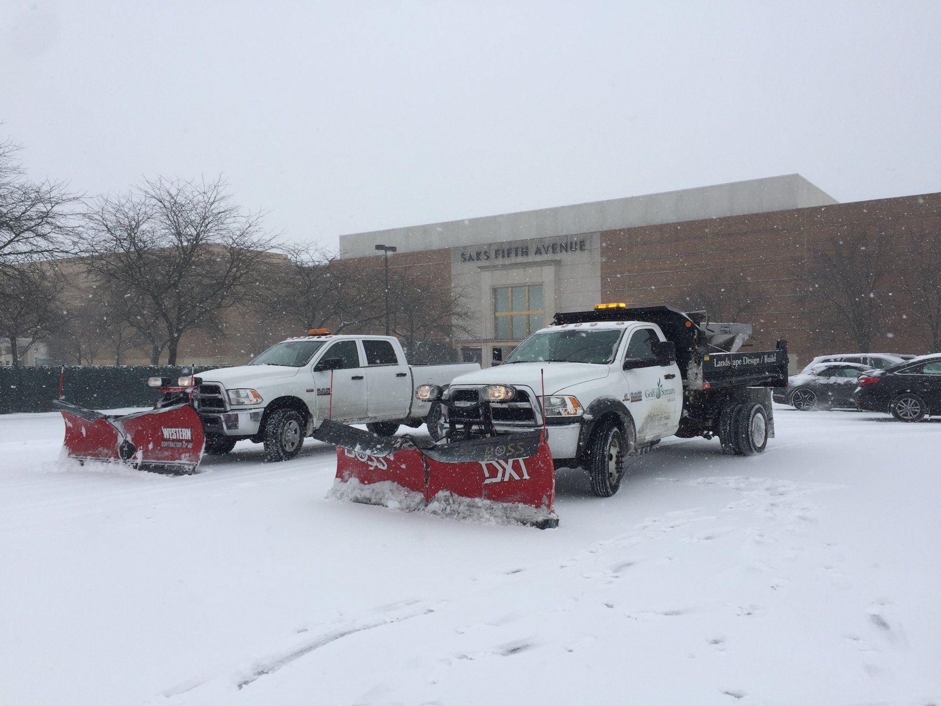 Two snow plows are parked in a parking lot in front of a building.